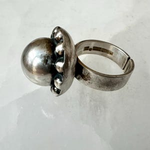 May include: A silver ring featuring a large, spherical central element and a textured, openwork border. The ring's band is thick and adjustable. The ring has a vintage, tarnished appearance.