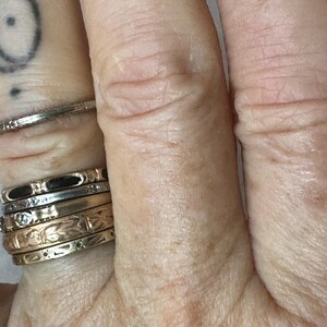 May include: Close-up of a hand adorned with multiple rings. The rings vary in style, including thin bands, engraved designs, and those with small gemstones. The hand also has a tattoo on the index finger.