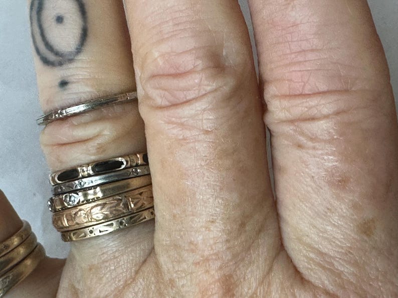 May include: Close-up of a hand adorned with multiple rings. The rings vary in style, including thin bands, engraved designs, and those with small gemstones. The hand also has a tattoo on the index finger.