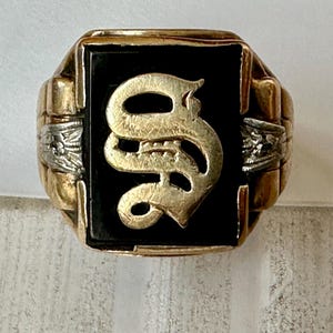 May include: A gold ring featuring a rectangular black onyx stone set in the center. A raised gold letter S in an old English font is mounted on the stone. The band is gold with silver-toned decorative side panels featuring etched details.