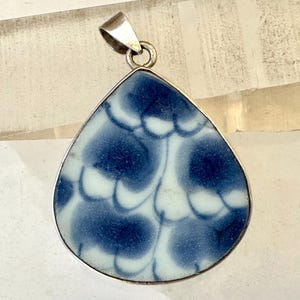 May include: A teardrop-shaped pendant with a silver bail. The pendant features a blue and white pattern, with dark blue shapes against a white background. The pendant is likely made of ceramic or glass, and is a piece of jewelry.
