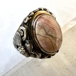 May include: A silver ring with an oval, pink and tan stone. The ring has ornate detailing around the stone and on the sides, including a seashell and fish design. The stone has a layered, marbled appearance.