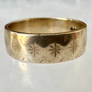 May include: A gold-colored ring with a wide band. The ring's surface is etched with a repeating pattern of starburst designs. The ring is displayed on a light-colored surface.