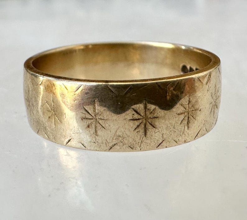 May include: A gold-colored ring with a wide band. The ring's surface is etched with a repeating pattern of starburst designs. The ring is displayed on a light-colored surface.