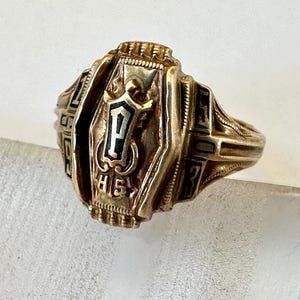 May include: A gold-colored class ring with black accents. The ring features the letter "P" inside a shield, with "HS" below. The ring has a detailed design and is likely a high school or college graduation ring.