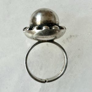 May include: A silver ring featuring a textured sphere atop a scalloped base. The adjustable band is visible. The metal has a slightly aged appearance. The ring is set against a light background. The design suggests a vintage or antique style, suitable for jewellery.