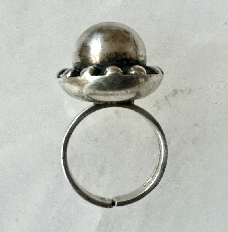 May include: A silver ring featuring a textured sphere atop a scalloped base. The adjustable band is visible. The metal has a slightly aged appearance. The ring is set against a light background. The design suggests a vintage or antique style, suitable for jewellery.