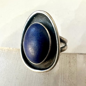 May include: A silver-toned ring featuring a large, oval, deep blue stone. The stone is set within a textured, open-backed frame. The ring has a double-band design for the finger.