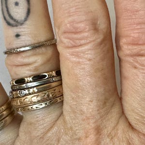 May include: Close-up of a hand adorned with several rings. The rings vary in style, including a thin, textured band, and others with floral and geometric designs. The rings are gold and silver in color.