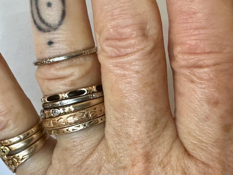 May include: Close-up of a hand adorned with several rings. The rings vary in style, including a thin, textured band, and others with floral and geometric designs. The rings are gold and silver in color.