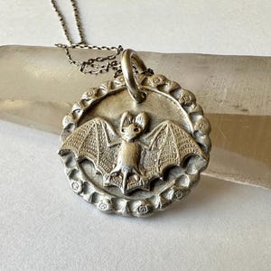 May include: Silver pendant necklace with a bat design. The bat is the focal point of a circular pendant with a textured edge, hanging from a fine chain. The pendant is approximately 3.8 cm in diameter.