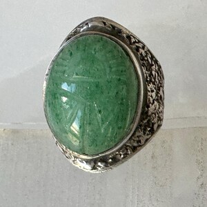 May include: A silver ring with a large, oval, green stone. The stone has etched details and is set in a textured silver band. The ring's design is vintage and eye-catching, suitable for everyday wear or special occasions.