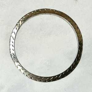 May include: A silver-toned ring with a repeating etched pattern. The ring's surface has a textured appearance, with a series of diagonal lines creating a visual texture. The ring is circular and appears to be made of metal.