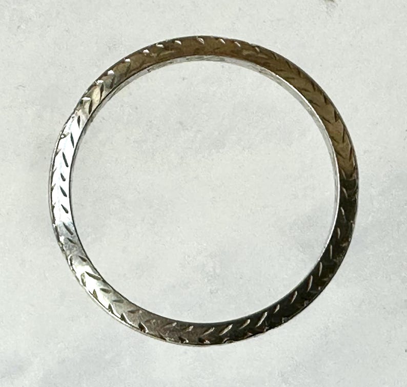 May include: A silver-toned ring with a repeating etched pattern. The ring's surface has a textured appearance, with a series of diagonal lines creating a visual texture. The ring is circular and appears to be made of metal.