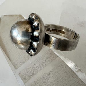 May include: A silver ring featuring a large, spherical, textured centrepiece. The ring's band is thick and plain, with hallmarks visible inside. The centrepiece has a distinctive, organic design, suggesting a vintage or antique piece.