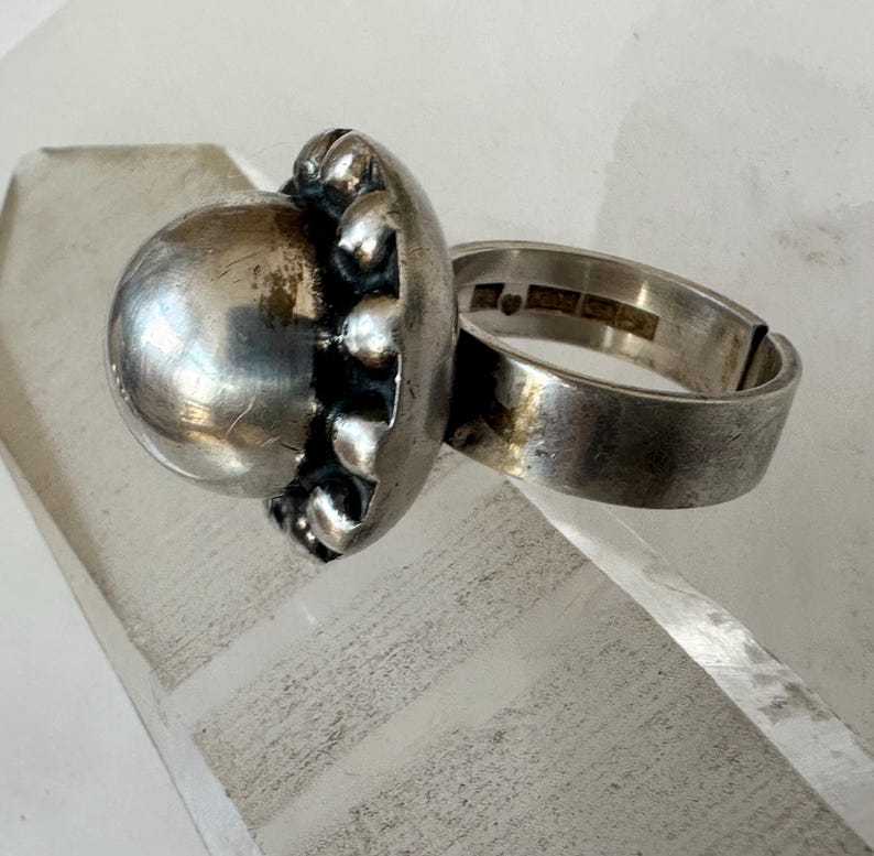 May include: A silver ring featuring a large, spherical, textured centrepiece. The ring's band is thick and plain, with hallmarks visible inside. The centrepiece has a distinctive, organic design, suggesting a vintage or antique piece.