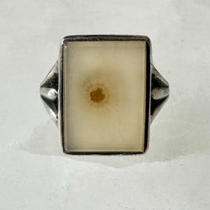 May include: A silver ring with a rectangular, cream-colored stone. The stone has a small, brown, flower-like design in the center. The ring's band has a simple, classic design.