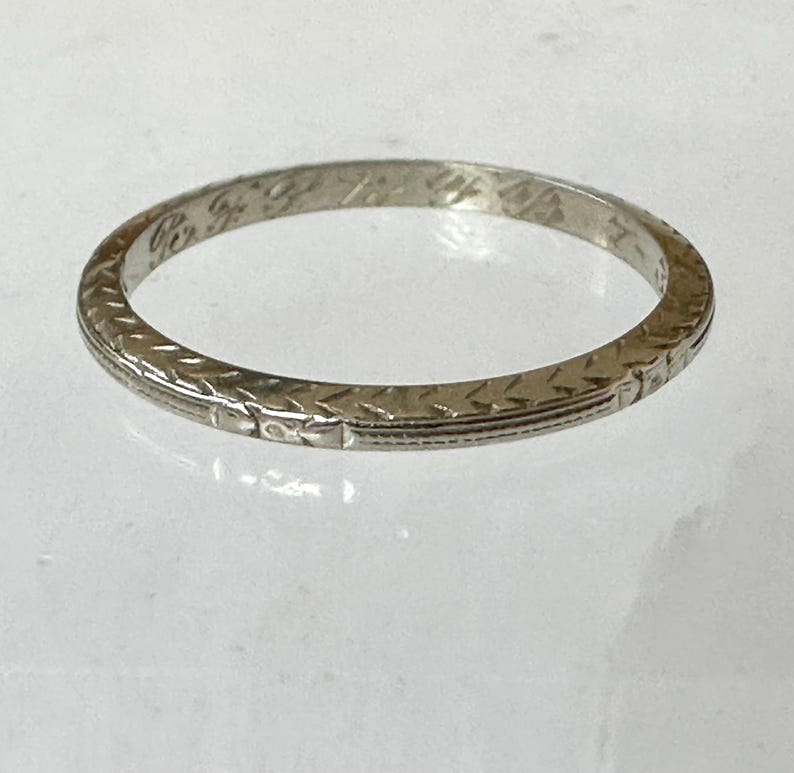 May include: A silver-colored ring with an engraved pattern. The ring has a repeating geometric design around the band. The ring appears to be made of metal and is likely a wedding band or a similar type of jewelry.