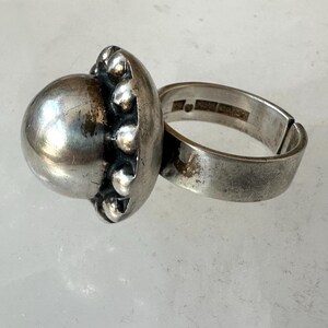 May include: A silver ring featuring a large, spherical central design and a textured border. The ring's band is broad and adjustable. The ring has a vintage or antique aesthetic.