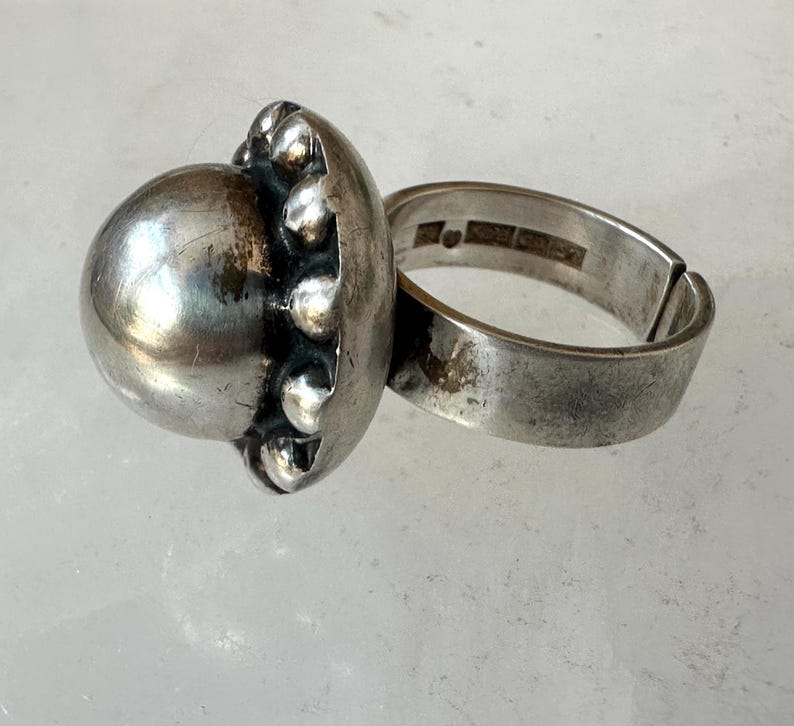 May include: A silver ring featuring a large, spherical central design and a textured border. The ring's band is broad and adjustable. The ring has a vintage or antique aesthetic.