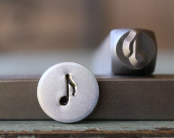 May include: A close-up of a metal stamp with a musical note design. The stamp is used to create impressions on metal.