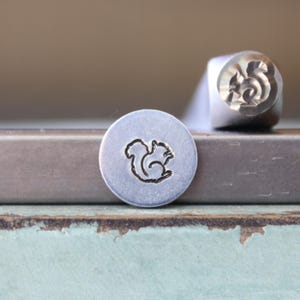Puede incluir: Un sello de metal con un diseño de ardilla. El sello está hecho de metal de color plateado y presenta un contorno detallado de una ardilla. Un círculo de metal estampado con el diseño de la ardilla está al lado del sello.