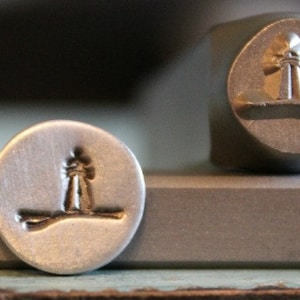 May include: Close-up of two metal stamps. One stamp is round with a lighthouse design. The other stamp is a block with a lighthouse design. The stamps are silver in color and are on a gray surface.