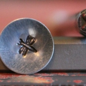 May include: A close-up of a metal stamp with a dragonfly design. The stamp is used to create impressions on metal.