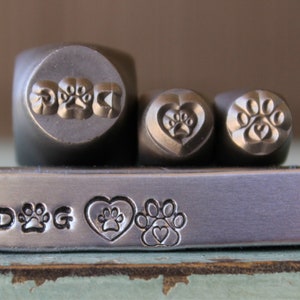 Puede incluir: Tres sellos de metal con diferentes diseños: una huella de pata, un corazón con una huella de pata y una huella de pata con un corazón. Los sellos se utilizan para crear joyas personalizadas u otras artesanías de metal. La palabra "DOG" está estampada en una barra de metal.
