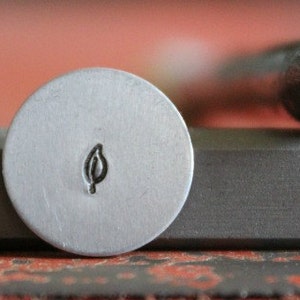 May include: A close-up of a metal stamp with a leaf design. The stamp is silver and has a round head. The leaf design is in the center of the head.