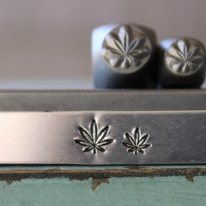 May include: A silver metal bar with two small leaf designs stamped into the surface. The bar is sitting on top of a green surface.