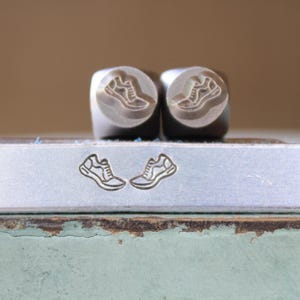 May include: Two metal stamps with a sneaker design, along with an impression of the same design. The stamps are cylindrical with a rounded top. The design is etched into the metal, showing the outline of a running shoe.