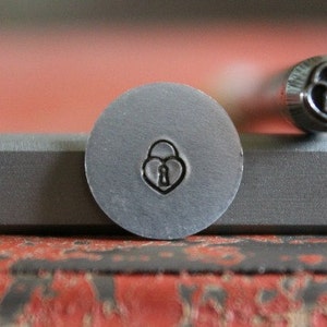May include: A close-up of a metal stamp with a heart-shaped padlock design. The stamp is silver and has a smooth, polished finish.