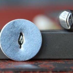 May include: A close-up of a metal stamp with a leaf design. The stamp is shown next to a metal surface with an impression of the leaf design.