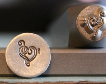May include: A close-up of a metal stamp with a design of a treble clef and heart. The stamp is used to create impressions on metal.