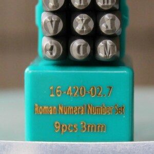 May include: A set of nine 3mm Roman numeral stamps in a turquoise plastic case. The stamps are made of metal and have a silver finish. The case has the text "16-420-02.7 Roman Numeral Number Set 9pcs 3mm" printed on it.