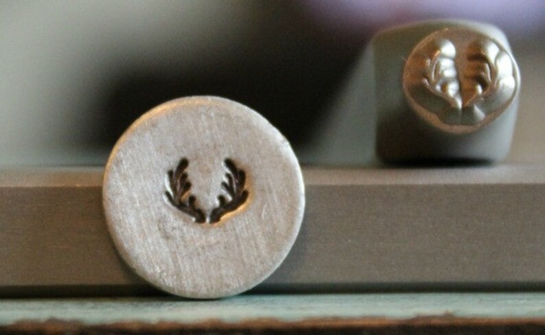 May include: A close-up of a metal stamp with a design of a wreath of leaves. The stamp is round and silver in color. The design is in the center of the stamp and is black in color.