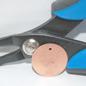 May include: A close-up of a black and blue metal hand punch tool with a copper disc being punched. The tool has a metal pin that is being pushed through the disc to create a hole.