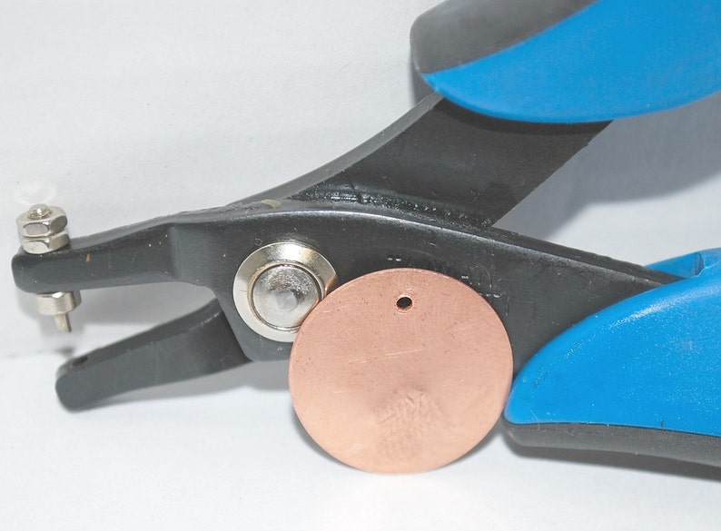 May include: A close-up of a black and blue metal hand punch tool with a copper disc being punched. The tool has a metal pin that is being pushed through the disc to create a hole.