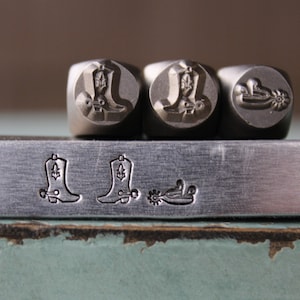 Peut inclure: Trois tampons métalliques avec des motifs de bottes de cowboy et une éperon. Les tampons sont disposés en rangée sur une barre en métal argenté. La barre est sur une surface bleue et verte.