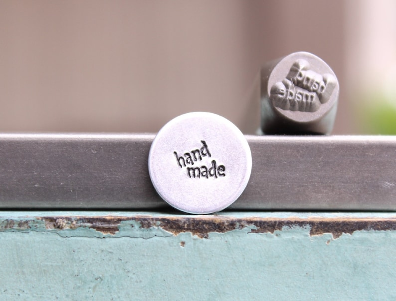 May include: A silver metal stamp with the words "hand made" engraved on it. The stamp is sitting on a silver metal surface.