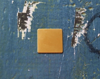 May include: A square, gold-colored metal plate on a blue, textured surface.