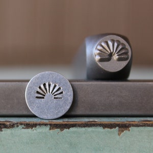 May include: A close-up of a metal stamp with a sunburst design. The stamp is used for creating impressions on metal.