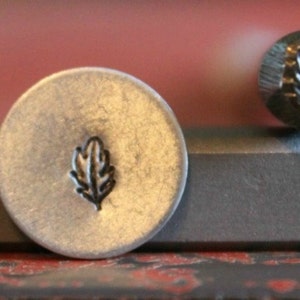 May include: A close-up of a metal leaf stamp. The stamp is used to create impressions on metal, leather, or other materials. The leaf design is simple and elegant.