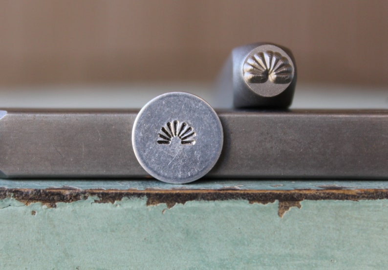 Pu&ograve; includere: Un set di timbri in metallo con un design a raggiera. Il set include un timbro rotondo con l'immagine della raggiera e un utensile per timbri abbinato. Gli utensili sono argentati e poggiano su una superficie metallica.