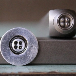 May include: A close-up of a metal stamp with a four-hole button design. The stamp is used to create impressions in metal.