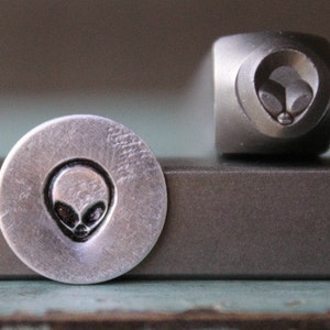 May include: A silver metal stamp with a design of an alien head. The stamp is used to create impressions on metal.
