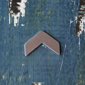 May include: A small, silver-colored metal chevron. The arrow-shaped piece has a matte finish and is set against a distressed, blue-green wooden surface. The chevron's design is simple and modern.