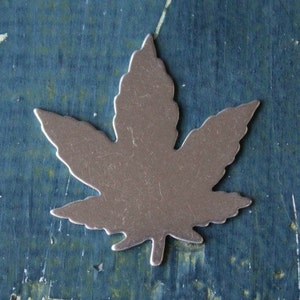 May include: A silver metal leaf charm in the shape of a marijuana leaf. The charm is on a blue background.