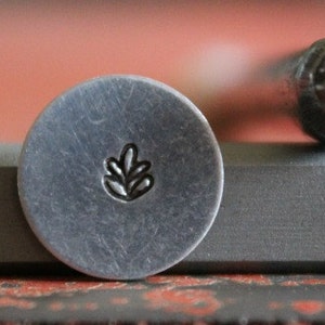 May include: A close-up of a metal stamp with a leaf design. The stamp is used for creating impressions in metal.